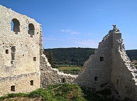 Links steht noch eine komplette Wand mit drei Rundbögenfenstern. Rechts sind nur noch die Teile zweier Mauern an der Ecke zu sehen. Im Hintergrund hat man einen schönen Blick ins Tal. Der Himmel ist wolkenlos.