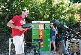 Pärchen am Ladeschrank Zwei Radler stehen am Ladeschrank an der Hammermühle. Die Frau in sommerlicher Kleidung hat gerade ein Ladefach geöffnet. Der Mann im roten Shirt hat bereits den Akku seines E-Bikes in der Hand.