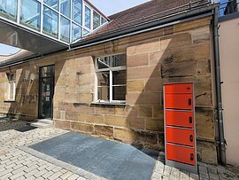 E-Bike und E-Rollstuhl-Ladestation Ein Haus mit brauner Steinfassade. Davor ein Schrnk mit roten Schließfächern. Zum Dach des einstöckigen Hauses führt ein verglaster Gang.