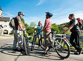 Vier E-Bike Fahrer stehen mit ihren Fahrrädern auf einem geteerten Radweg und unterhalten sich. Im Hintergund stehen Häuser im fränkischen Baustil.