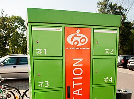 Ein Ladestation für elektronische Fahrräder. Ein in etwa 1,5 Meter hoher, grüner Metallkasten mit acht einzelnen Schließfächern. aus orangenem Hintergrund steht in großen Buchstaben das Wort Ladestation.