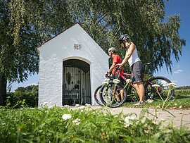 Zwei Radler, eine Frau mit weißem Shirt und ein Mann in rot, halten mit ihren Fahrrädern vor der Kapelle Warching. Die Kapelle steht unter einem großen Baum.