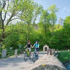 Ein Pärchen ist mit ihren Fahrrädern auf dem Radweg unterwegs. Rechts begleitet sie die Anlauter. Der Wald strahlt in hellem grün. Hinter ihnen sieht man eine kleine Kapelle.