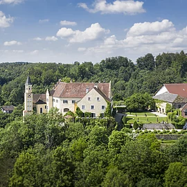 Das Schloss Hexenagger von oben zu sehen. Eingebettet in grünem Blätterkleid. Der Himmel ist hellblau und weiße Wolken sind zu sehen.