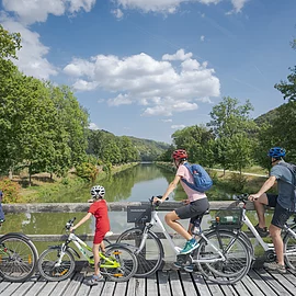 Eine Familie mit dem Rad unterwegs hält an einer Brücke am Ludwig-Donau-Main-Kanal bei Altessing. Das Ufer ist von grünen Bäumen gesäumt und der Himmel strahlt hellblau.