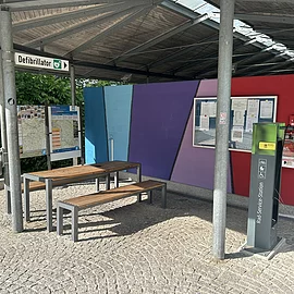 Unter dem Dach am Großparkplatz steht eine Radreparaturstation mit der man die nötigsten Reparaturen an seinem Fahrrad vornehmen kann. Links ist ein Tisch und zwei Bänke für eine kleine Rast.