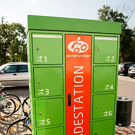 Ein Ladestation für elektronische Fahrräder. Ein in etwa 1,5 Meter hoher, grüner Metallkasten mit acht einzelnen Schließfächern. aus orangenem Hintergrund steht in großen Buchstaben das Wort Ladestation.