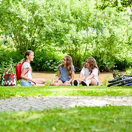 Drei junge Mädchen sitzen auf einer Wiese am Ufer eines Flusses. Sie unterhalten sich. Rechts von ihnen liegt ein Fahrrad. Im Hintergund fließt ein Fluss. Am gegenüber liegenden Ufer wachsen Bäume und Sträucher.