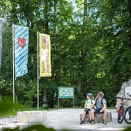 Zwei Radler sitzen auf einem Felsbrocken am Geographischem Mittelpunkt. Rechts von ihnen sind ihre zwei Fahrräder abgestellt. Hinter ihnen ist rechts eine Infotafel und links von ihnen sind drei Fahnen gehisst.