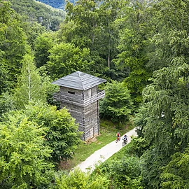 Der Limesturm aus Holz inmitten eines Waldes, der hellgrün leuchtet. Der Limesradweg läuft direkt am Turm vorbei. Hier sind zwei Radler auszumachen.