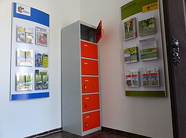 Ein orange-grauer Indoor-Ladeschrank für eBike-Akkus mit insgesamt sechs Fächern und einem offenen Fach steht zwischen zwei Wanddisplays mit Broschüren in dem Vorraum der Touristinformation Monheim.