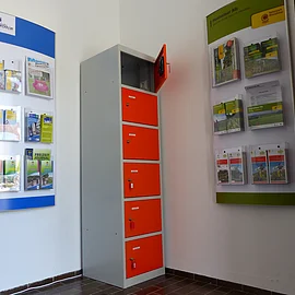 Ein orange-grauer Indoor-Ladeschrank für eBike-Akkus mit insgesamt sechs Fächern und einem offenen Fach steht zwischen zwei Wanddisplays mit Broschüren in dem Vorraum der Touristinformation Monheim.