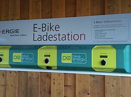 Ladestation E-Bike-Ladestation mit drei Ladepunkten an einer Holzwand. Schild mit Logo und Informationen.