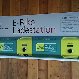 Ladestation E-Bike-Ladestation mit drei Ladepunkten an einer Holzwand. Schild mit Logo und Informationen.