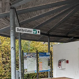 Perfekt für eine Pause Überdachter Rastplatz mit Holzbänken, Infotafeln und einem Defibrillator an der Wand.