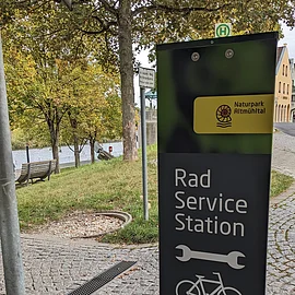Hilfe für kleine Reparaturen Fahrradservicestation mit Werkzeug- und Fahrradsymbol, im Hintergrund Bäume und ein Fluss.