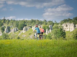 Radfahrer bei den 12 Apostel Solnhofen Zwei Personen fahren mit Fahrrädern auf einem grasbewachsenen Feld vor einer Felslandschaft.