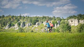 Zwei Personen fahren mit Fahrrädern auf einem grasbewachsenen Feld vor einer Felslandschaft.