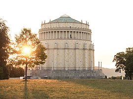 Befreiungshalle in Kelheim von Fotograf Anton Mirwald Rundes historisches Gebäude mit Statuen auf dem Sims, Sonne hinter Baum im Park bei Abendlicht