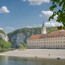 Flussufer mit Kiesstrand, altes Gebäude mit Turm und bewaldete Felsen unter blauem Himmel mit Wolken