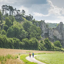 Zwei Radfahrer auf einem Weg vor bewaldeten Felsen mit einem Kreuz auf dem höchsten Felsen.