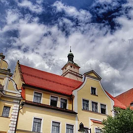 Historische gelbe Geb&auml;ude mit roten D&auml;chern und Kirchturm unter bew&ouml;lktem Himmel.