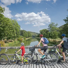 Vier Personen mit Fahrrädern auf einer Holzbrücke mit Blick auf einen Fluss und grüne Landschaft.