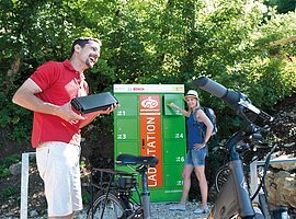 Mann h&auml;lt Fahrradakku, Frau &ouml;ffnet gr&uuml;nen Fahrrad-Ladestation-Schrank im Freien bei Sonnenschein